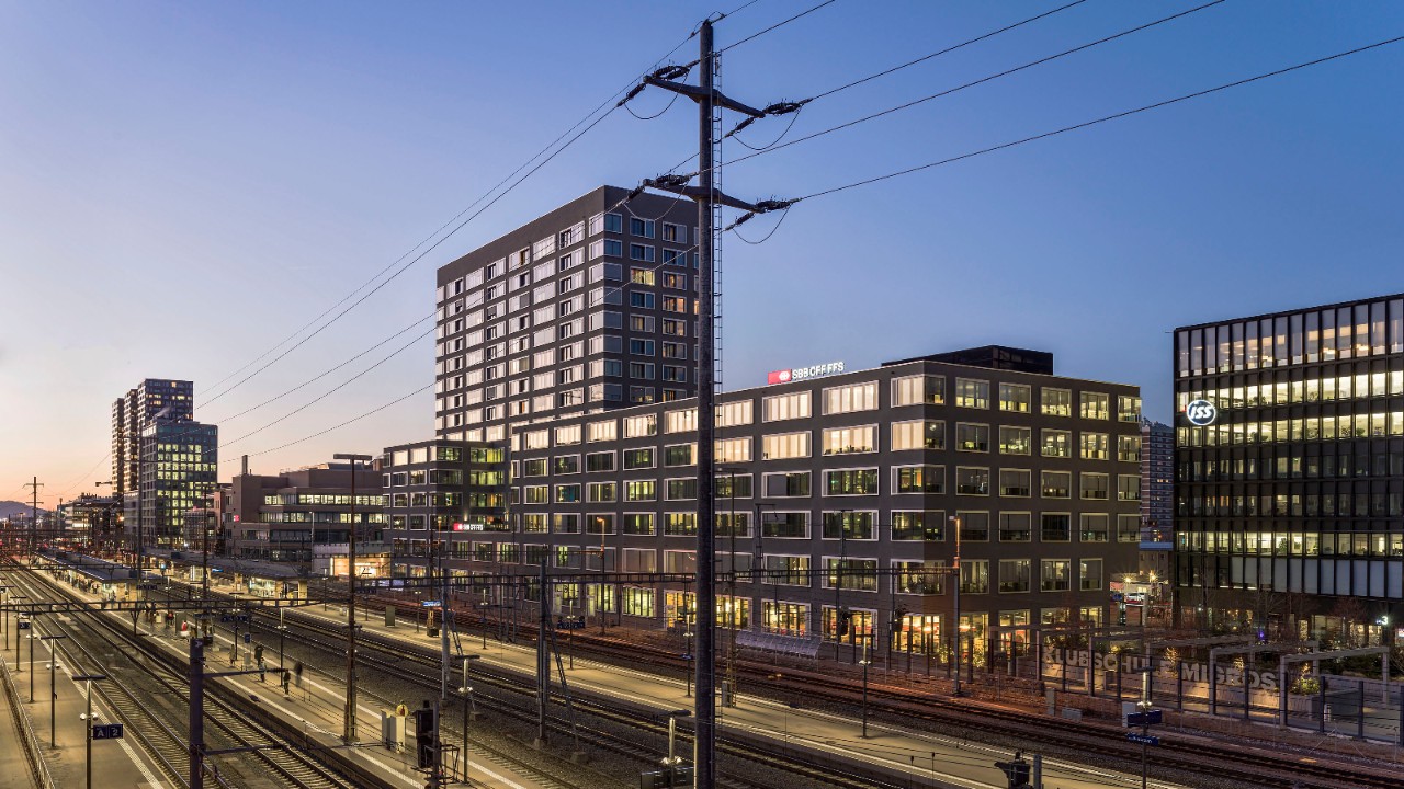 L’image montre plusieurs immeubles de bureaux éclairés à côté d’une gare au crépuscule.