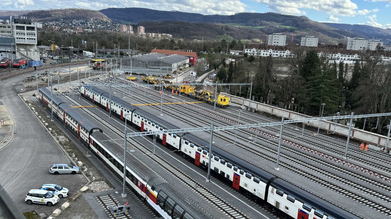 Luftaufnahme einer grossen Abstellanlage mit mehreren parallel stehenden Zügen auf Gleisen. Im Hintergrund sind gelbe Wartungsfahrzeuge und Gebäude sichtbar, dahinter bewaldete Hügel und Wohnhäuser. Links im Bild befinden sich ein Parkplatz und Zufahrtsstrassen.