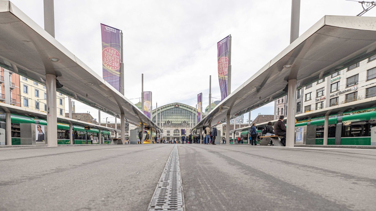Der Centralbahnplatz in Basel ist belebt mit Passanten und grünen Trams unter einer modernen, geschwungenen Überdachung. Bunte Banner fügen dem grauen Betonplatz Farbe hinzu, während im Hintergrund das historische Gebäude des Bahnhofs Basel SBB thront. Der Himmel ist bedeckt, was dem gesamten Platz eine weiche Beleuchtung verleiht.