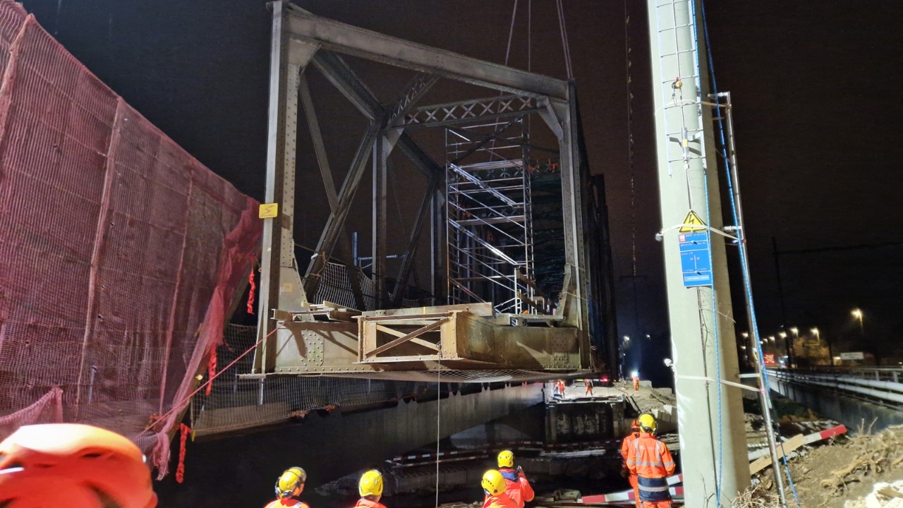 Das Bild zeigt wie ein massives Stahlfachwerk einer Brücke aus seiner ursprünglichen Position ausgehoben wird. Die Szene ist durch künstliches Licht beleuchtet, der Fluss ist erkennbar. 