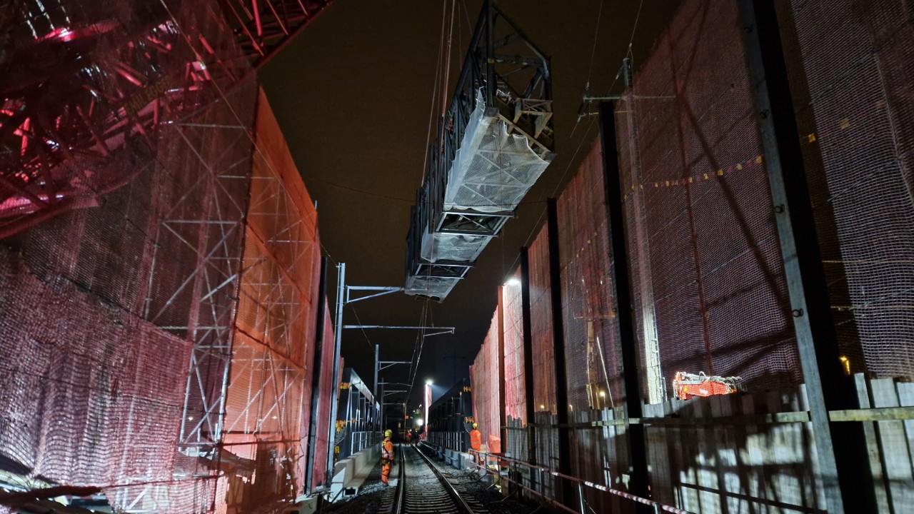 Das Bild zeigt wie der Kran das Stahlfachwerk der alten Brücke über die neue Brücke hebt. Die Szene ist durch künstliches Licht beleuchtet. Die neue Brücke ist durch temporäre, rote Schutzzäune flankiert. 