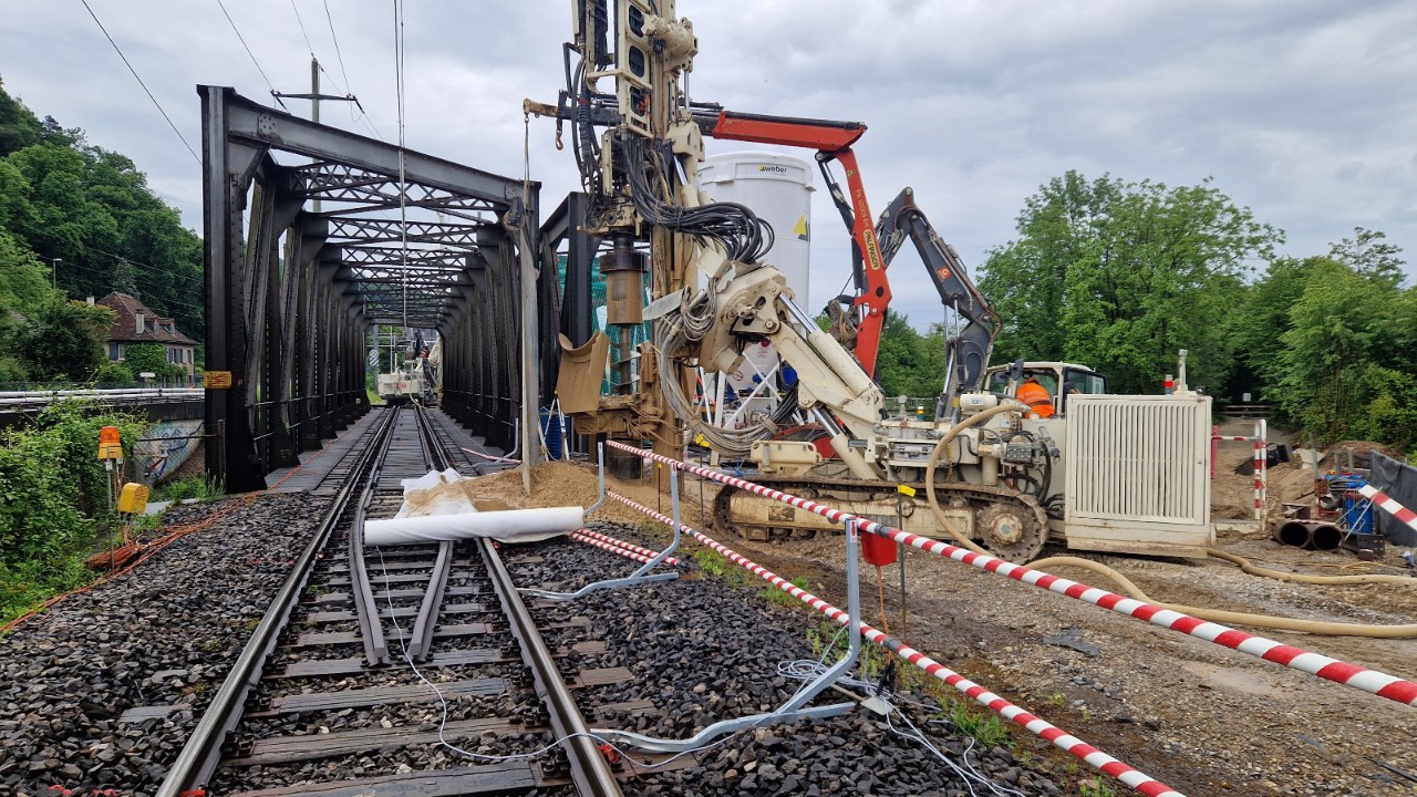 Das Foto vom 1. Juni 2024 zeigt die Vorbereitungsarbeiten zum Aushub der oberen Birsbrücke. Die Bahnstrecke war an diesem Wochenende aus Sicherheitsgründen komplett gesperrt. Das Bohrgerät rechts im Bild erstellt Rühlwandträger als Abschluss der künftigen Baugrube für die neuen Brückenwiderlager. Gleichzeitig wird sodann das Bauschutzgerüst auf diesen Trägern montiert. Links im Bild, am Ende der unteren Birsbrücke, ist ein Bagger und das andere Bohrgerät erkennbar. Sie nehmen auf der anderen Flussseite dieselben Arbeiten vor. 