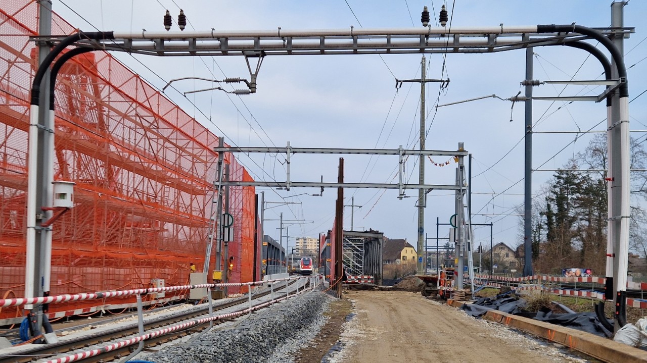 Zwei Brücken liegen nebeneinander. Links sieht man das Stahlfachwerk der neuen Brücke, auf welche gerade ein Zug zugefahren kommt. Rechts liegt das Stahlfachwerk der alten Bahnbrücke, zu welcher keine Gleise mehr führen. 