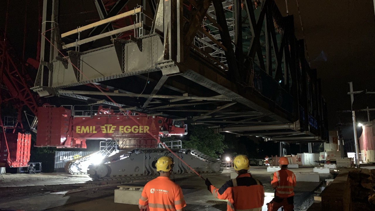 Das Foto vom 19. Juni 2024 zeigt, wie der Raupenkran das Stahlfachwerks auf den Installationsplatz heruntersenkt. Die Bauleute sorgen mit Seilen dafür, dass sich das Stahlfachwerk in der Luft in der richtigen Bahn bewegt und nicht zu schwenken beginnt. Auf dem Installationsplatz wird das Stahlfachwerk in den folgenden Tagen zerlegt, abtransportiert und fachgerecht entsorgt.