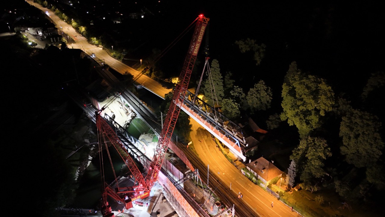 Bei Nacht hebt ein großer roter Kran ein Stahlfachwerk über eine Straße und setzt es präzise auf die Pfeiler einer Bahnbrücke. Die Baustelle ist hell ausgeleuchtet, um die Arbeiten im Dunkeln zu ermöglichen. Das Fachwerk bildet ein zentrales Element der neuen Brückenkonstruktion. 