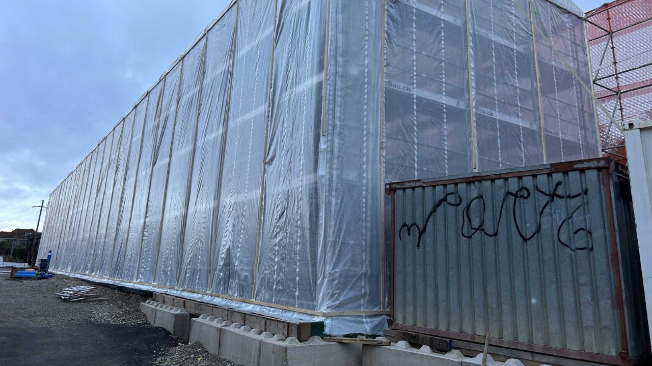 Das Foto zeigt eine fertiggestellte, transparente Einhausung, die das darunterliegende Stahlfachwerk während der Beschichtungsarbeiten schützt. Neben der Einhausung ist ein Container mit sichtbarem Graffiti zu erkennen. Die gesamte Struktur ruht auf einem Betonfundament und ist von Bauzäunen umgeben, während der Himmel im Hintergrund bewölkt erscheint.