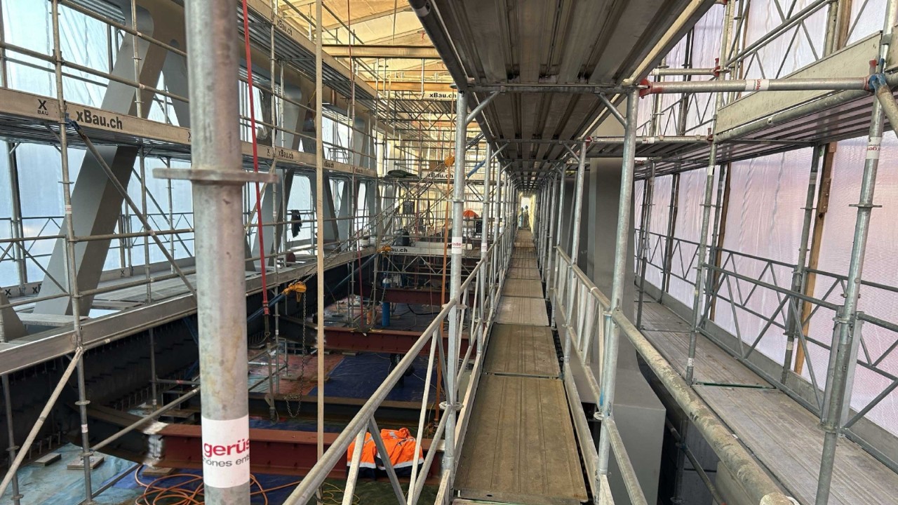 Das Foto zeigt das Innere einer Einhausung, in der ein Bauteam beschäftigt ist. Umgeben von Gerüsten und Ausrüstungen, tragen die Arbeiter sorgfältig Beschichtungen auf. Die Komplexität und der Detailreichtum der Struktur betonen die Intensität ihrer Arbeit.
