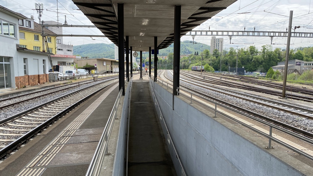 Das Foto zeigt den Mittelperron des Bahnhofs Olten Hammer mit Gleisen auf beiden Seiten und einer gedeckten Perrondachkonstruktion. Links sind das Bahnhofsgebäude und angrenzende Bauten sichtbar, rechts erstreckt sich der Formationsbahnhof mit mehreren Gleisen. Die Aufnahme blickt entlang des Perrons in Richtung Olten und zeigt das bestehende Bahnumfeld.