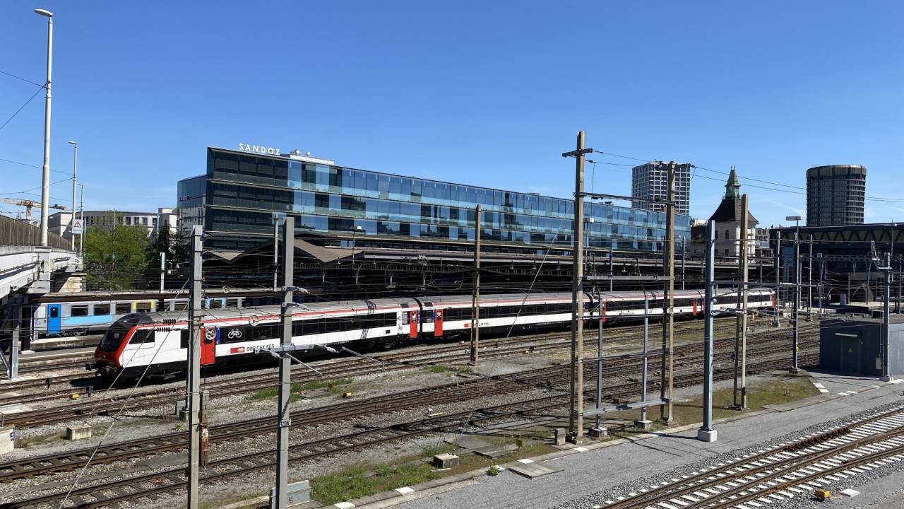 Blick über mehrere Gleise des Bahnhofs Basel SBB mit einem stehenden SBB-Zug im Vordergrund und dichtem Netz aus Fahrleitungen und Masten. Dahinter stehen die Perronhalle des Bahnhofs SNCF sowie das moderne Gebäude «Elsässertor» mit spiegelnder Glasfassade; links verläuft die Margarethenbrücke. Rechts sind weitere Perrons sichtbar, die im Rahmen des Projekts erweitert werden sollen.